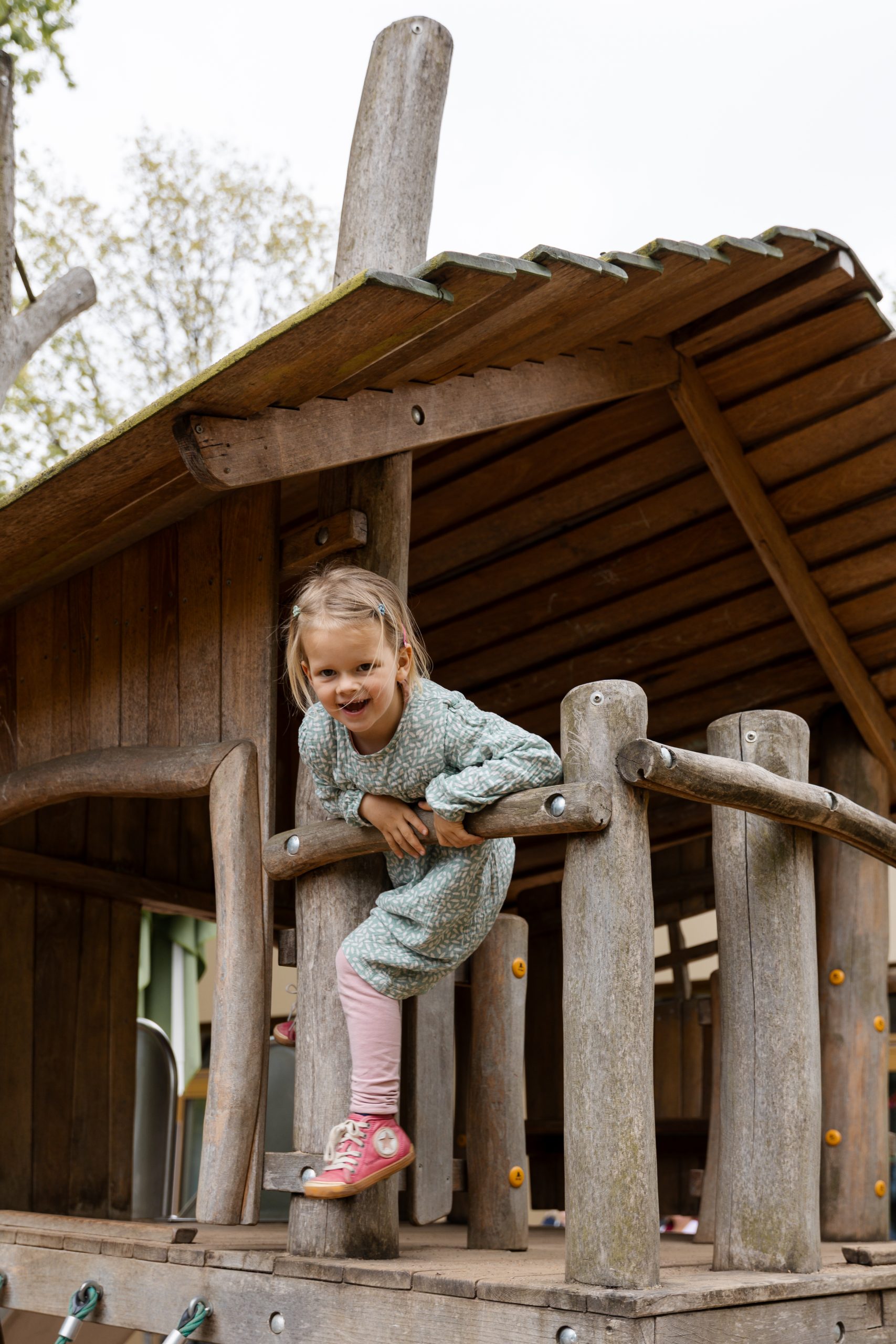 Mädchen klettert hoch auf dem Baumhaus und schaut fröhlich zur Kamera – natürliche Kitafotografie Hamburg